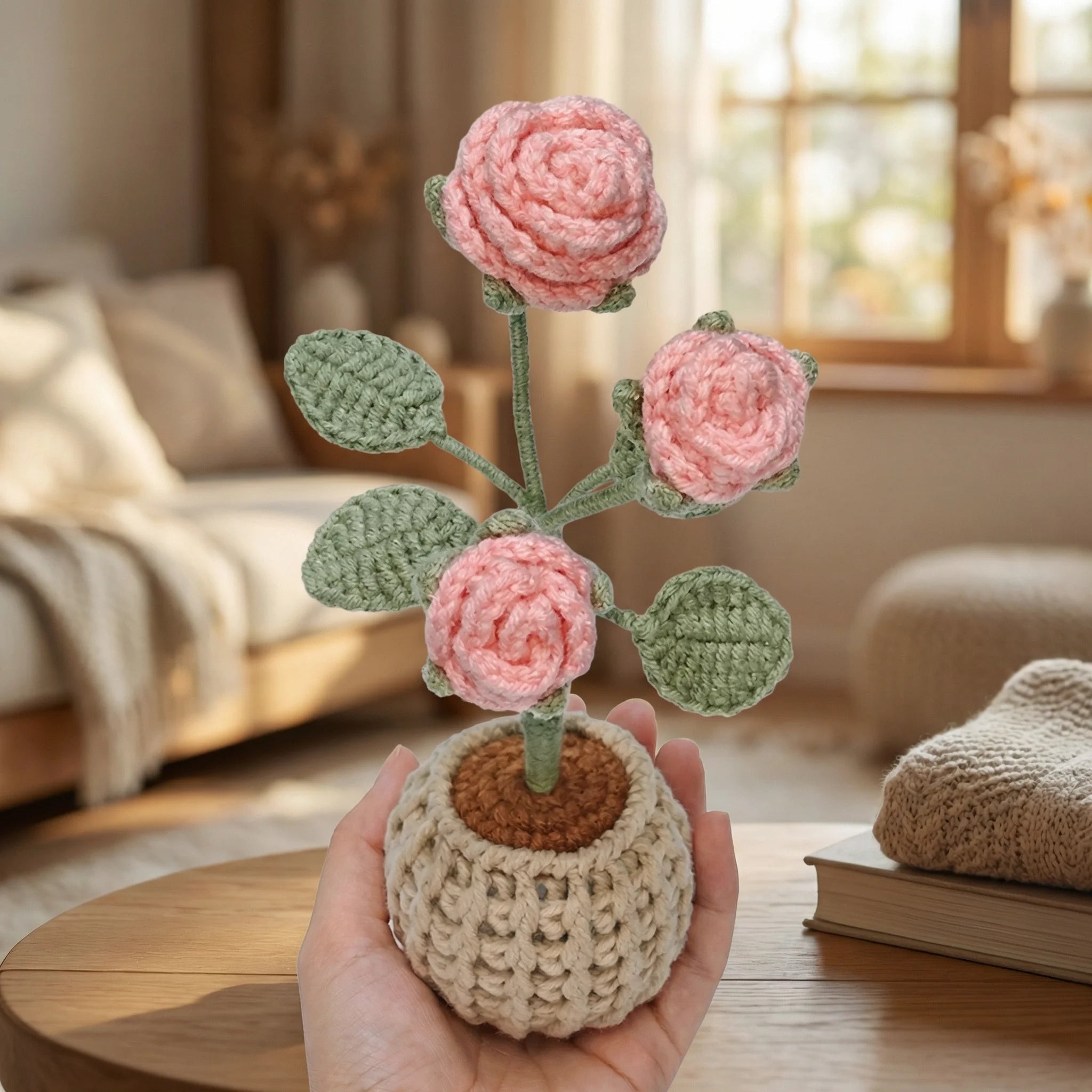 Hand holding The Rose Garden mini crochet kit with pink roses in beige basket pot on table with books