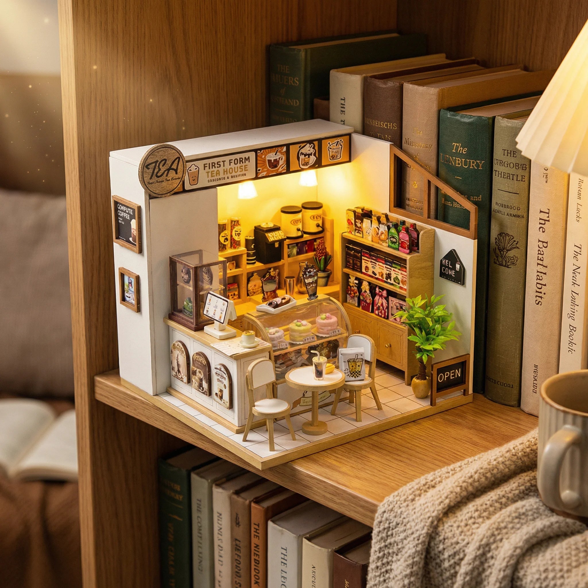 The Tea House miniature DIY kit displayed on bookshelf with warm lighting showing detailed coffee shop interior scene