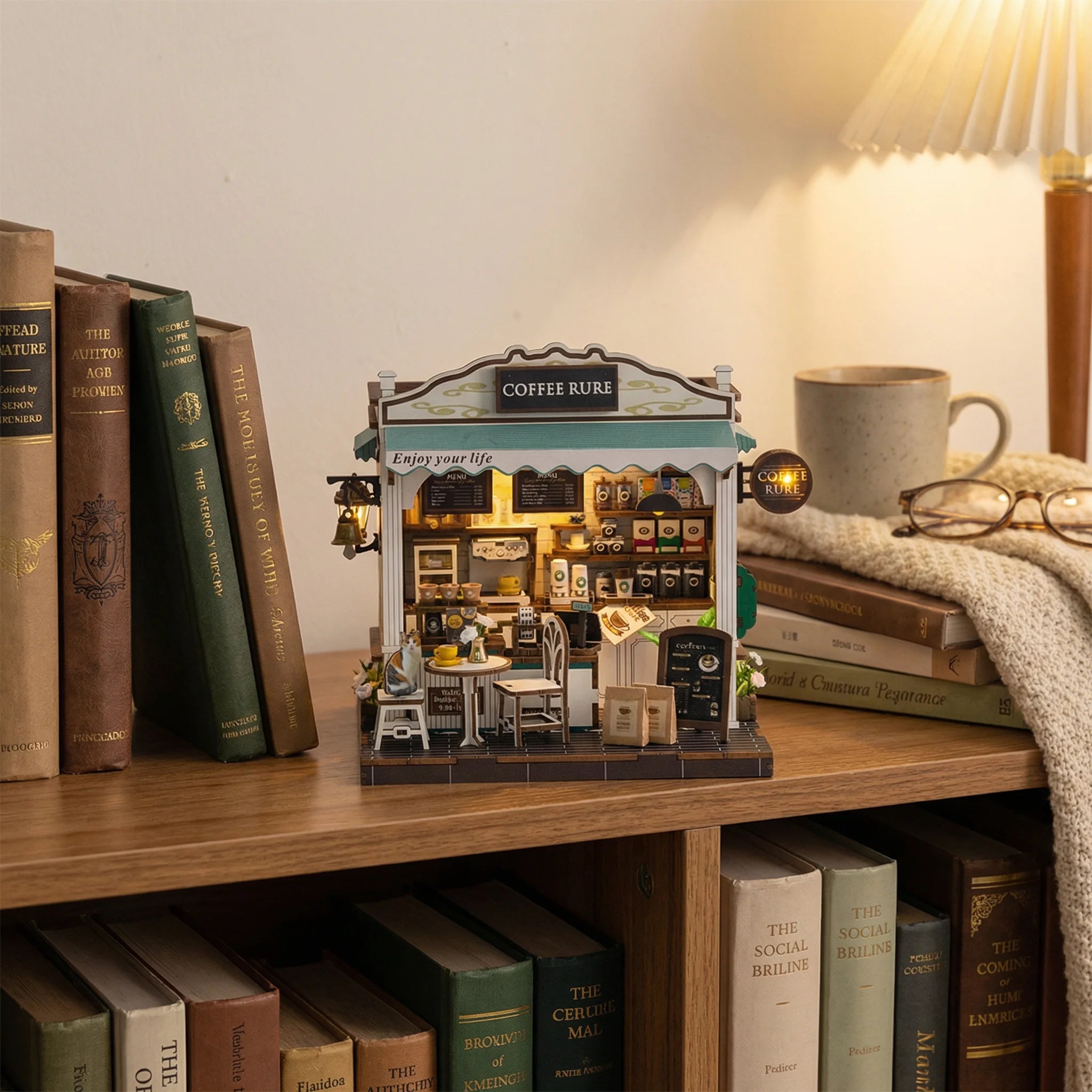 The Coffee Stop miniature DIY kit displayed on wooden shelf with books, lamp, coffee mug and cozy blanket