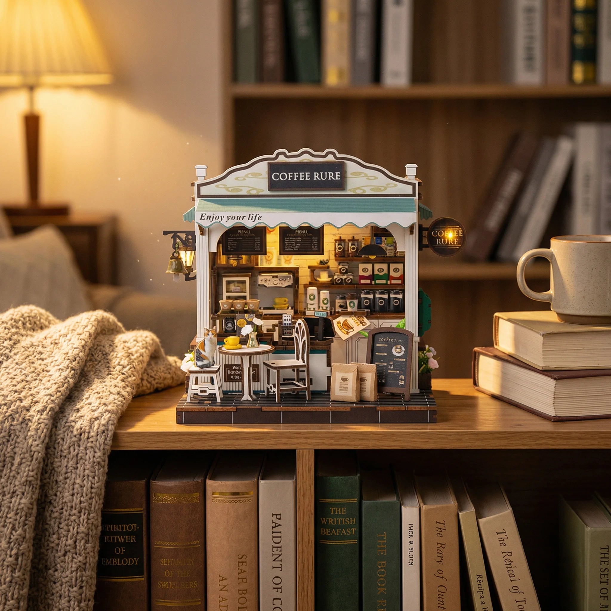 The Coffee Stop miniature DIY kit displayed on wooden shelf with books, lamp, and cozy blanket in home setting