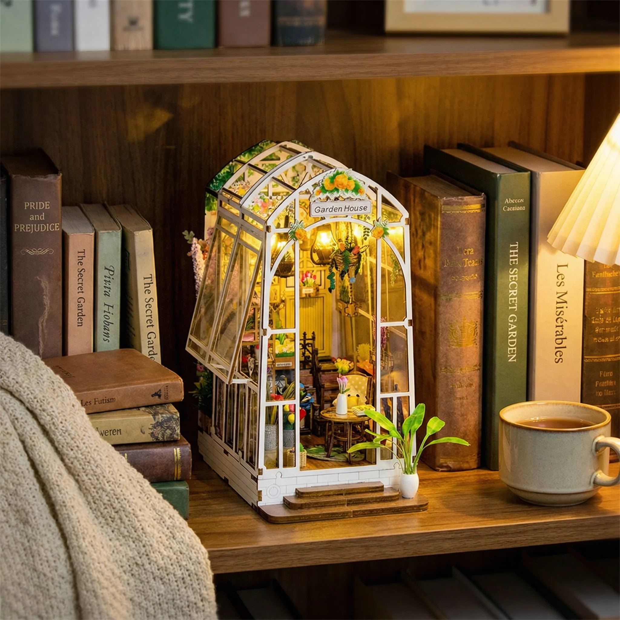 The Garden House miniature greenhouse kit displayed on bookshelf with warm lighting next to coffee mug and classic books