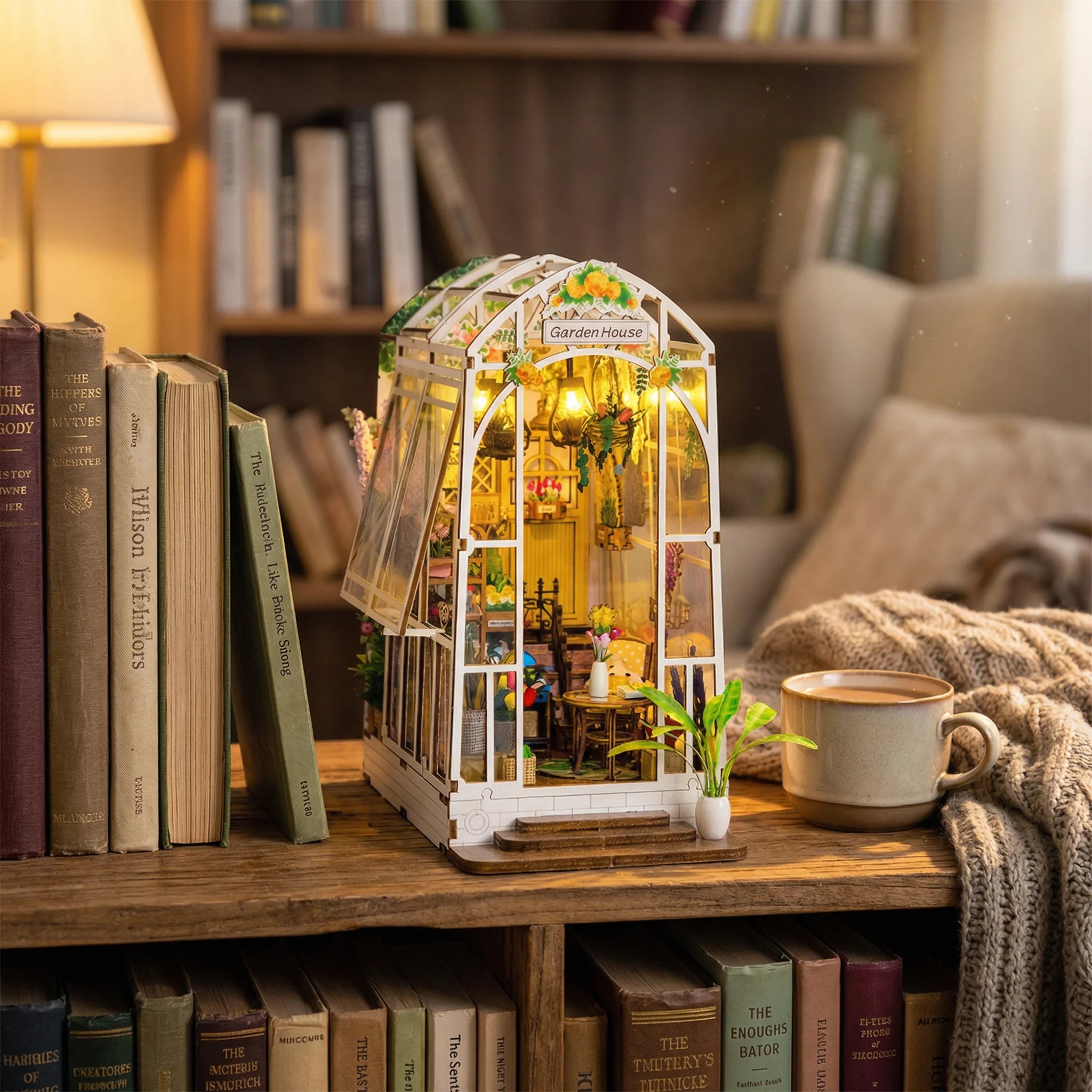 The Garden House miniature greenhouse kit displayed on wooden shelf with books, coffee mug, and cozy blanket