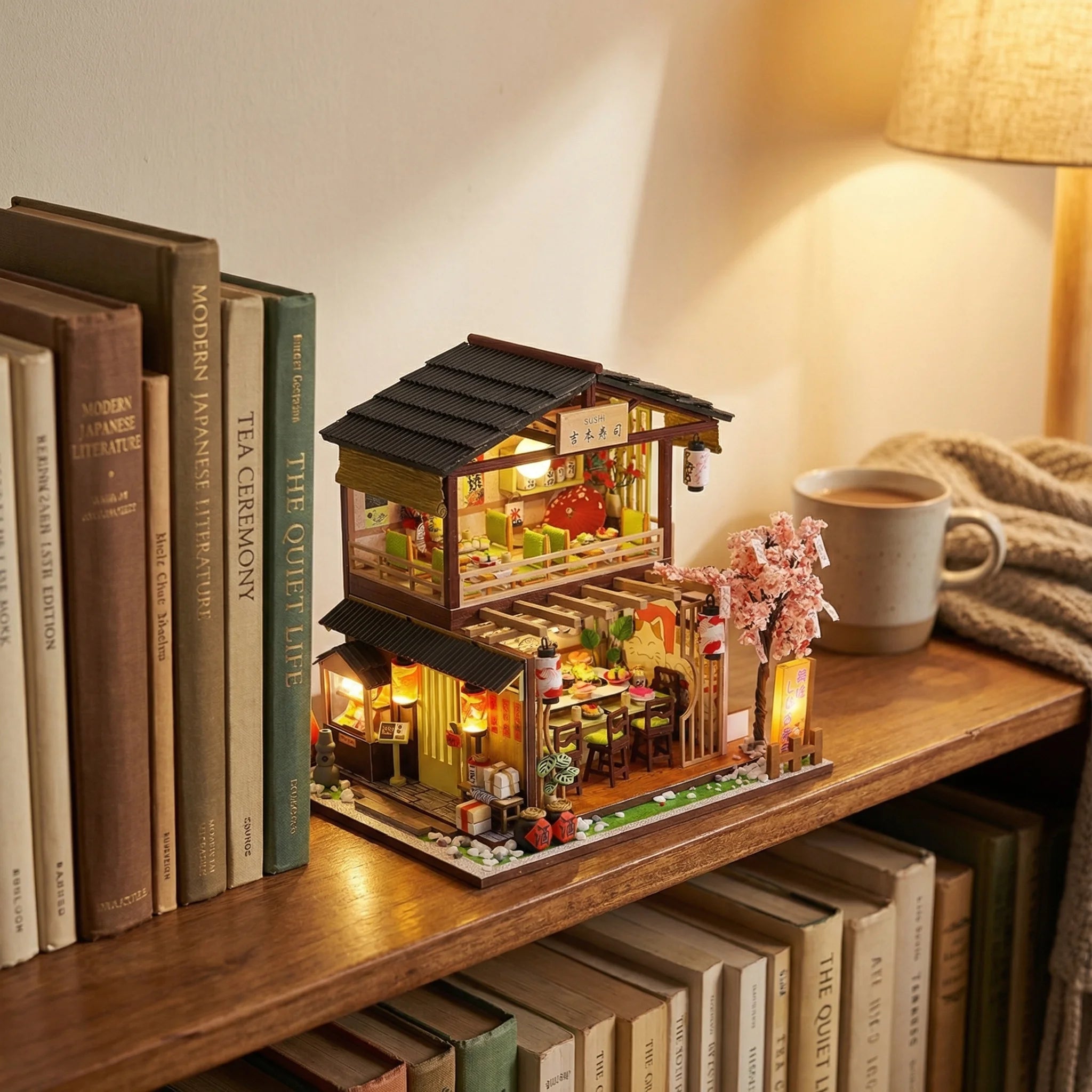 The Sakura Sushi Bar miniature dollhouse kit displayed on wooden bookshelf with warm lighting and cherry blossom tree