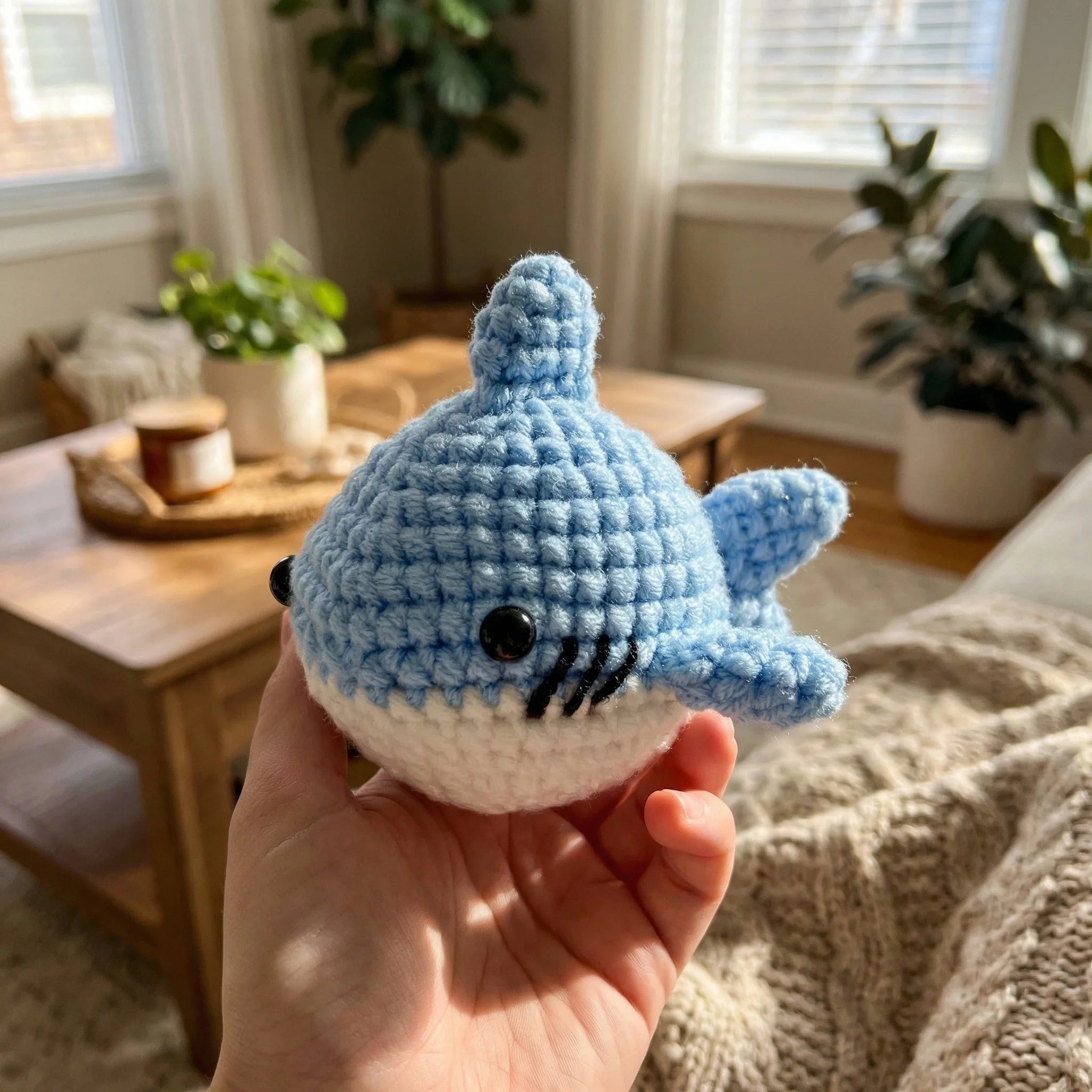 Hand holding The Ocean Grin mini crochet shark amigurumi in cozy living room with natural light