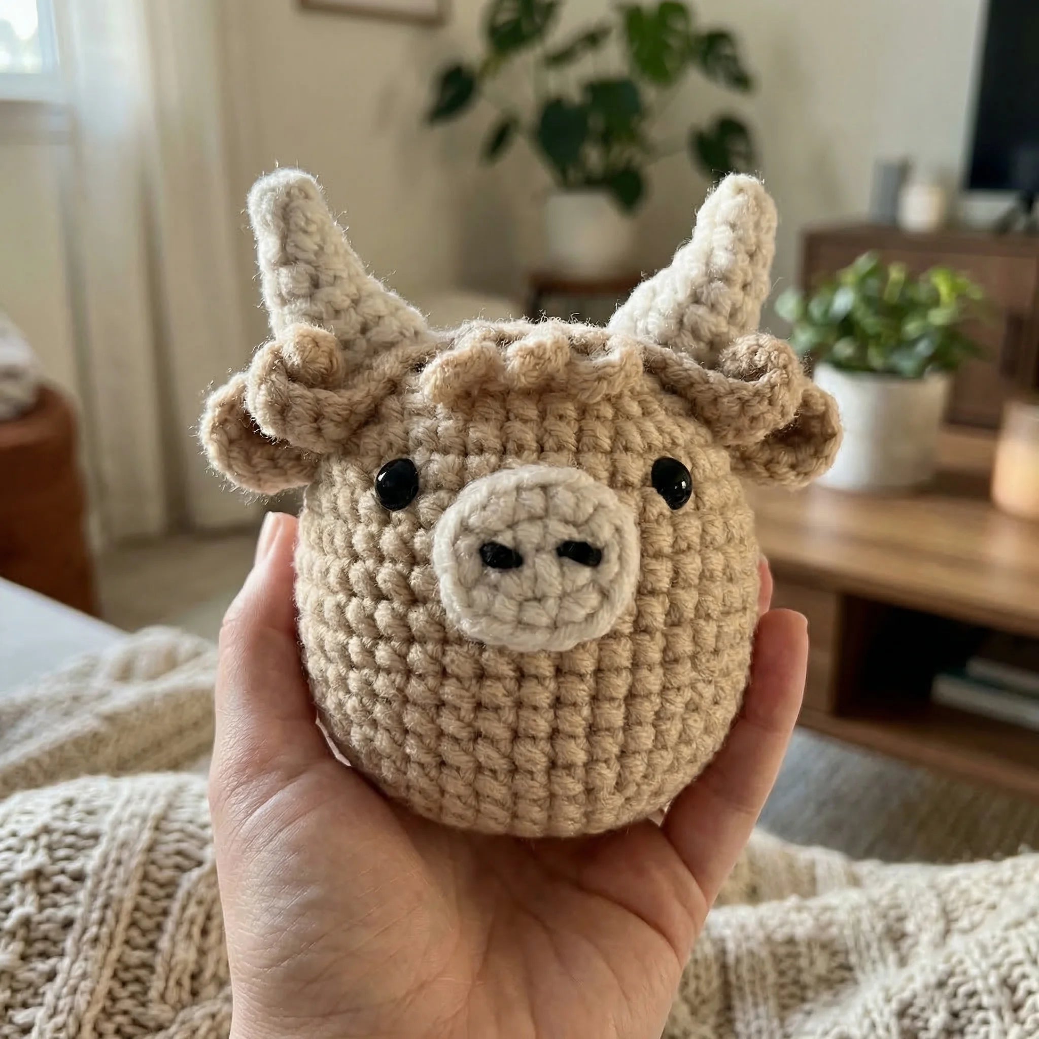 Hand holding Hamish the Highland Cow mini crochet amigurumi with beige fuzzy yarn texture in cozy home setting