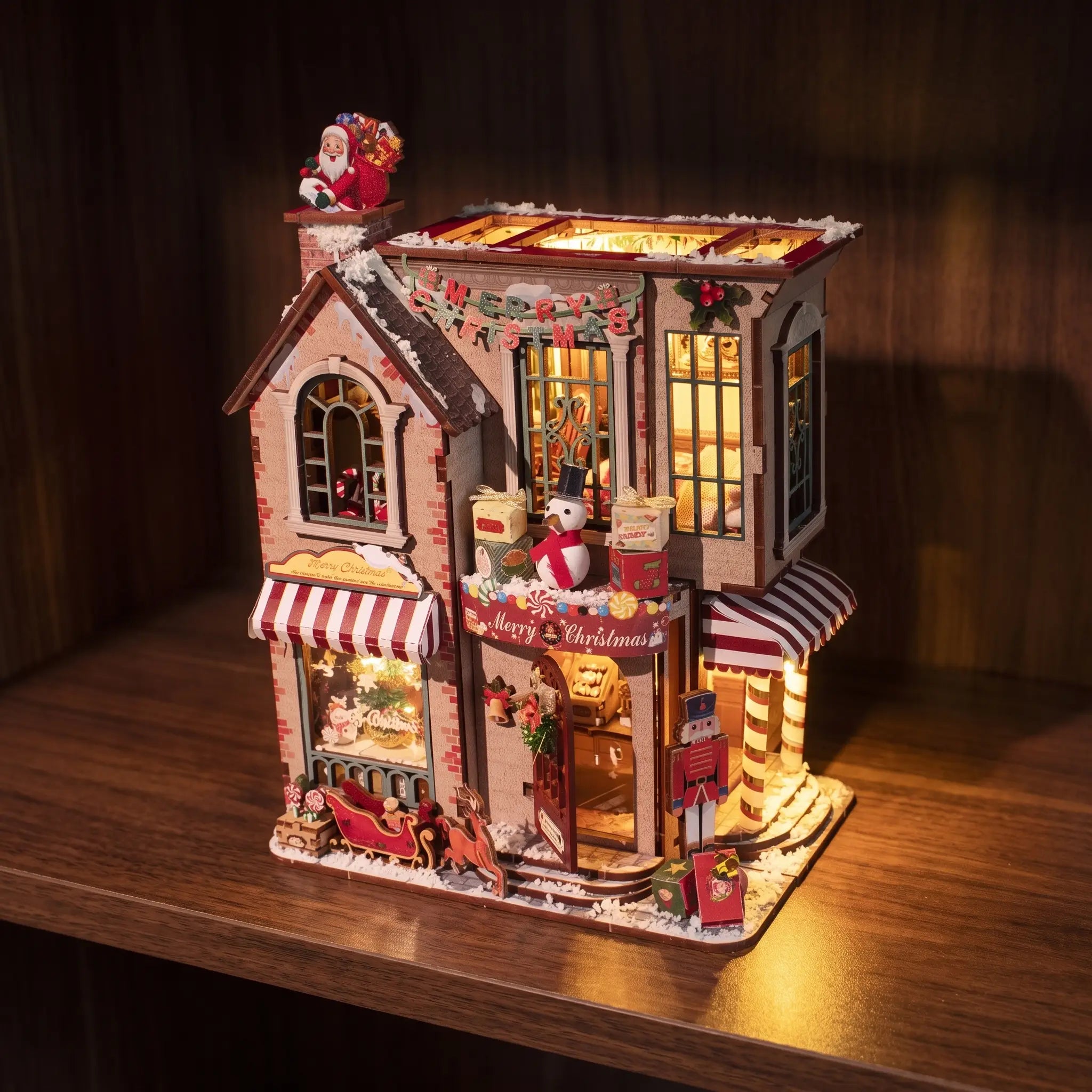 The Candy Cane Cottage miniature kit glowing on a dark walnut shelf at night